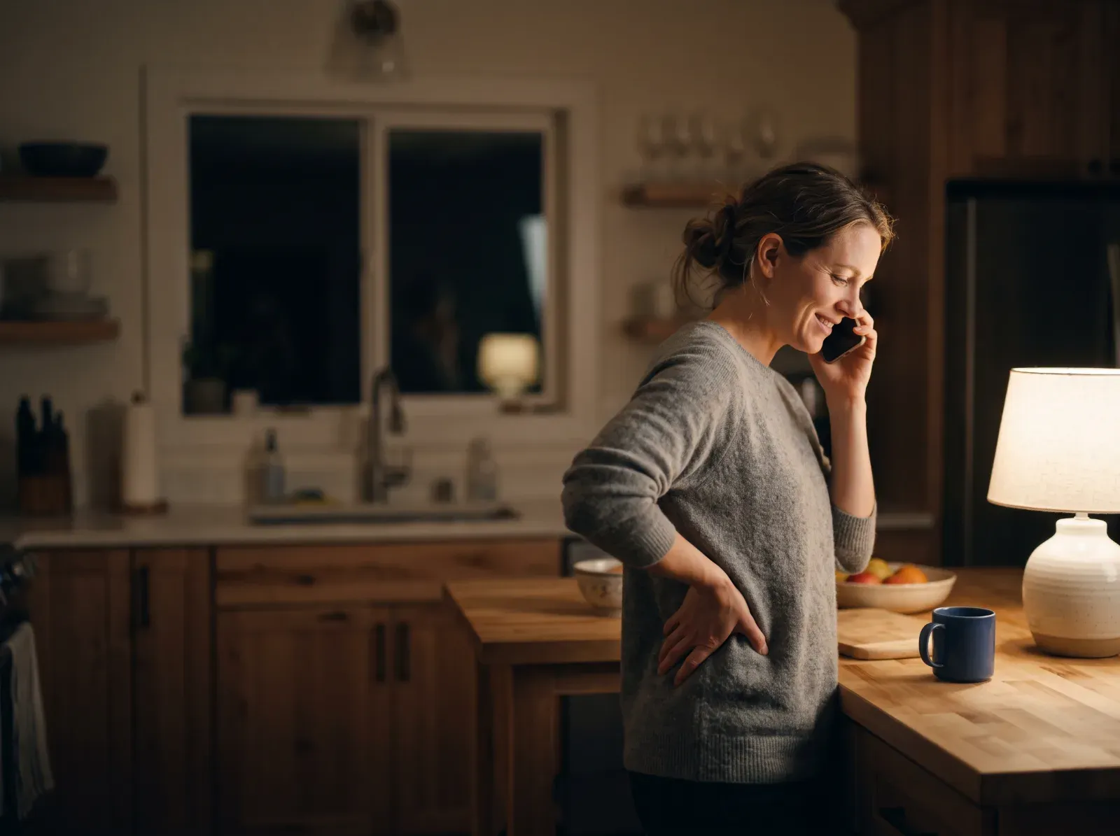 A woman in her kitchen on the phone, one hand on her lower back